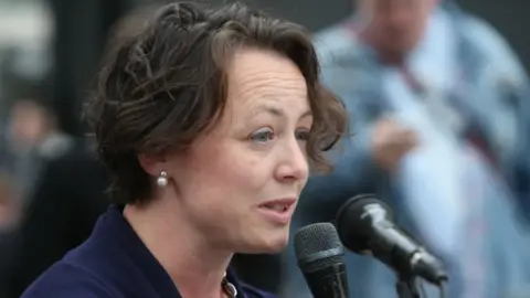 Catherine McKinnell, Labour MP for Newcastle, is talking into microphones. She has short dark hair and is pictured side on.