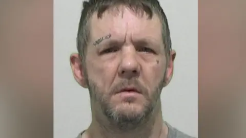 Mugshot of McKenna. He has a lined face, untidy short brown hair, a grey beard and tattooed symbols above his right eye and a heart tattooed on his left cheek.