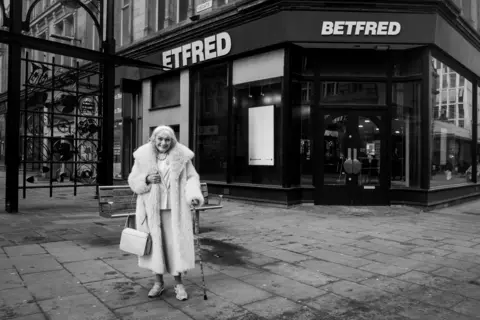 A black and white photo of an elderly woman standing outside a betting shop in Bradford city centre.