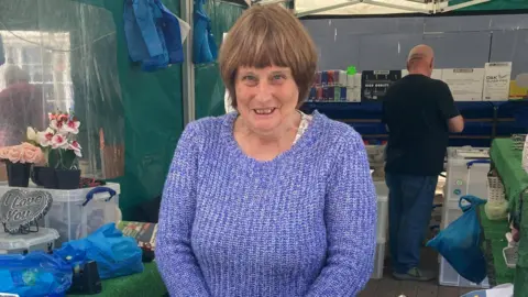 A woman standing in a market stall - in front of her are a series of ornaments of football badges, including Manchester United, West Bromwich Albion and Birmingham City. She is wearing a purple jumper, and behind her is another trader with his back turned.
