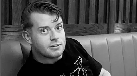 247 Kettles Black and white photo shows young man with black t-shirt on smiling while on soft seating