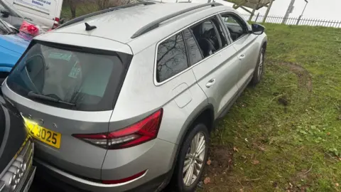 Máirtín Mac Gabhann A grey SUV sits diagonal across the bottom of a green grass slope. Its rear rests on a black car's grill. Their are brown skid marks on the slope and a black metal fence sits at the top of the slope. 