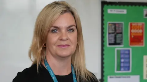 Shaun Whitmore/BBC A head and shoulders image of a woman. She is standing in a classroom. She is wearing a black top, golden hoop earrings and a blue lanyard around her neck. She is smiling and looking into the camera. 