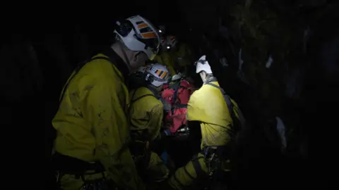Underground rescue team COMRU in yellow overalls surround a red-blanketed stretcher inside a cave