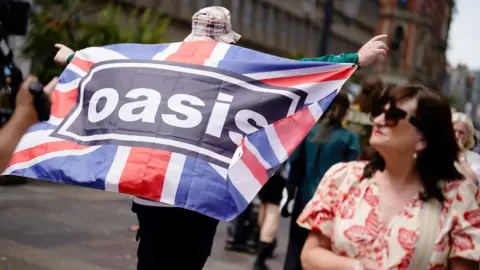 An Oasis fan in a bucket hat walking through the centre of Cardiff. He has a Union flag draped across his shoulder with the Oasis logo printed on it. He is walking away from the camera into a crowd of people. On the right hand side of the picture, in the foreground, is a woman with shoulder length dark hair wearing dark sunglasses and a cream dress with red printed leaves on it.