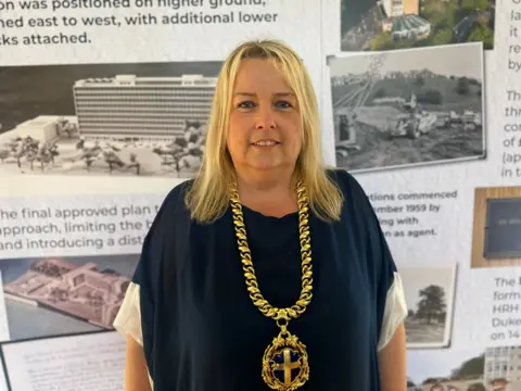 Amanda Hopgood smiles at the camera. She is wearing a large gold chain and medallion and has shoulder-length blonde hair.
