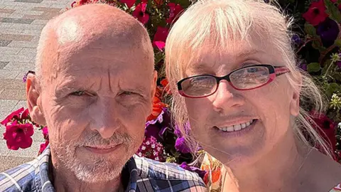 LDRS handout Close up of Ron who has white receding hair and white beard and wears a blue, white and checked shirt and Tracey who has white hair tied back and wears black and red glasses. They are pictured in front of flowers. They are both smiling. 