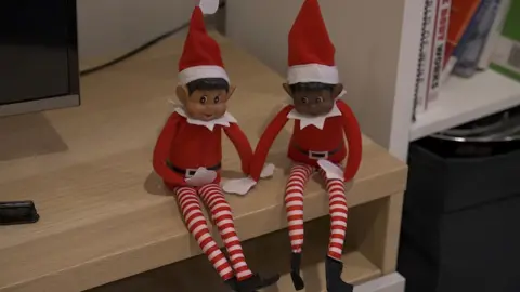 Two toy elves with red and white stripy tights sit on a wooden television stand with part of the TV visible.