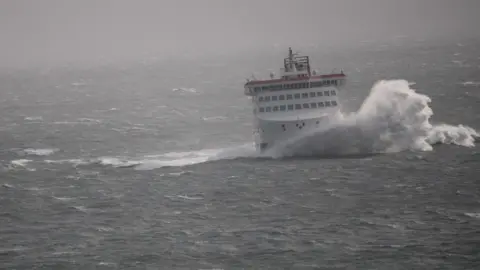 The Manxman ferry in stormy conditions. The white and red vessel is in choppy seas and a large white wave crashes in front of it.