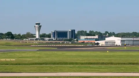 Runway at Birmingham Airport surrounded by grass. The airport buildings, including a control tower, can be seen in the distance against the green trees on the horizon.