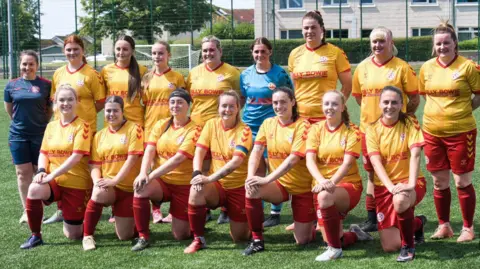 JazzaMcG Photography A team photo of a women's football team, wearing yellow shirts and red shorts.