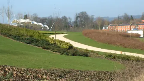 Maidstone Borough Council A park, with green lawns, a beige path running through it with a white structure to the left and a row of reddish houses to the right