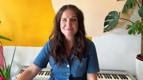 Amy Joce smiles at the camera as she sits in front of an electric keyboard and some plants. She is wearing a denim jumpsuit and has long brown hair.