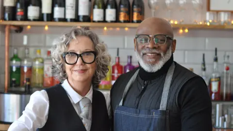 Jayne Casey, with grey curly hair and black rimmed glasses, wears a shirt, tie and black pinafore. Smiling, she stands next to a manwho is also wearing black rimmed glasses. Eric Gooden wears a black jacket and denim apron. They are both standing in front of a bar.