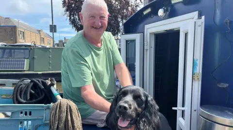 Olivia Richwald/BBC A man in a green T-shirt smiles at the camera. He is petting a spaniel.