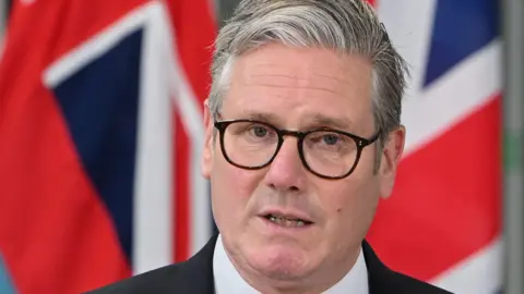 A close-up of Sir Keir Starmer's face, wearing glasses and pictured in front of a Union Jack flag, as he speaks to news media.