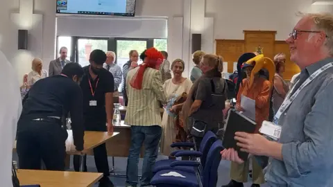Nadia Lincoln/LDRS A large number of people at a council meeting, showing a man wearing a jester hat and another man with a wig on his head. Two people, in black T-shirts are moving a table.