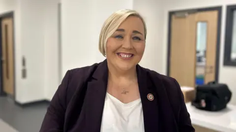 Fiona Kane. She has short blonde hair, wearing a thin silver necklace, white top and purple blazer.