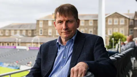 Gloucestershire County Cricket Club The chairman of Gloucestershire County Cricket Club Peter Matthews sits in the stands at the county ground in Bristol. He has dark hair and is wearing a blue jacket and blue shirt. The pitch is visible in the background