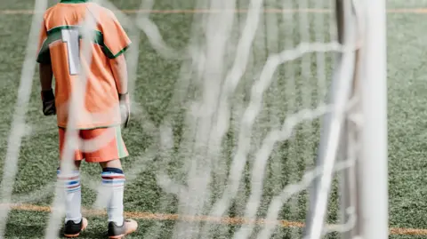 A photo taken through the net behind a goal showing a goalkeeper in an orange kit, wearing gloves and the number 1 jersey
