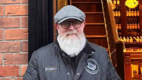 A man is standing infront of a shop door. He has a black coat and grey hat on. He is wearing glasses and has a large grey beard. The building behind him is made from red bricks. Inside the shop door there is a set of steps and a shop counter.