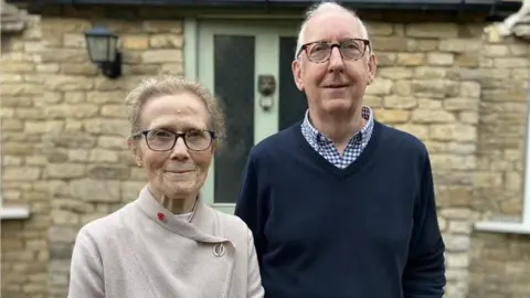 Alex Dunlop/BBC Marion and Tim Shepherd, standing outside a stone brick house. They are both looking at the camera, smiling. Marion, to the left is wearing a cream top, with a poppy at the neck, her hair tied back and is wearing glasses. Tim, next to her has short grey hair, wearing glasses, a checked shirt and blue jumper on. The house has a green front door and a light to the left. 