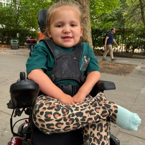 Charlie Brown A young blonde girl in a black wheelchair with black straps across her chest. She is wearing a green Wicked t shirt and leopard print trousers. Behind her is a pavement and green-leaved trees.