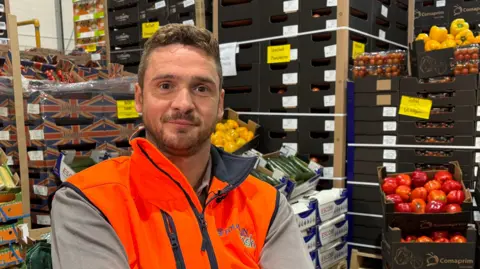 Ben Schofield/BBC Jack Hanson, wearing an orange high-visibility jacket and looking straight at the camera, smiling slightly. He has a short, dark beard, and mousy-coloured hair, worn short at the sides and slightly longer on top. He is seated in front of dozens of boxes of fresh produce, including red and yellow peppers, courgettes, tomatoes and sweetcorn. 