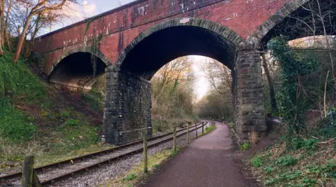 A section on the cycle path running parallel to a section of railway. There is also a bridge running above the cycle path. The path is lines by banks with trees and undergrowth.