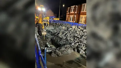 Northumbrian Water A yellow digger working in the dark on Ellen Terrace. There is a hole in the road exposing a water pipe underneath. It is surrounded by concrete debris and water. The area is fenced off with blue barriers.