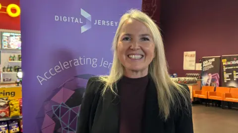 A woman called Katie King smiles broadly at the camera. She is stood in front of a purple Digital Jersey sign. She has long blonde hair which is straight and reaching past her shoulders. She has a dark purple top on and a black jacket.