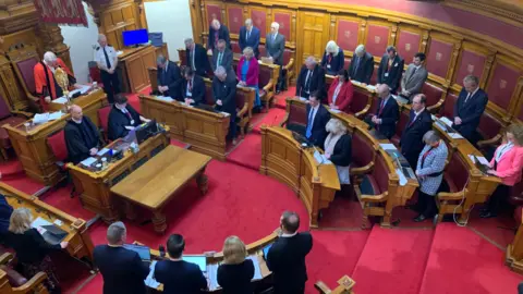 BBC The States Assembly with smartly dressed men and women standing around the chamber with heads bowed. There are wooden panelled walls and desks and a bright red carpet