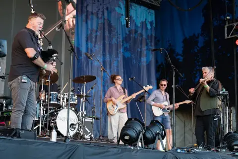Getty Images A traditional music band performs on a stage in front of several microphones, including a male bagpipe player, a drummer who is obscured, a female bass player, a male guitarist and a male fiddle player