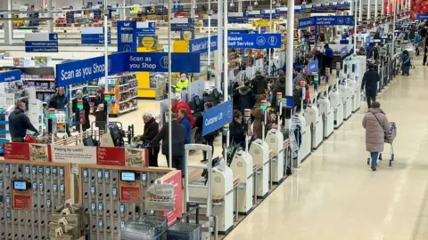 Gambar getty orang berbelanja di dalam tesco besar dengan pemindaian saat Anda berbelanja tanda dan pemindai dan tanda-tanda swalayan terlihat