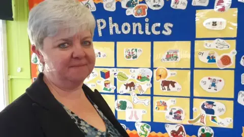 Sarah Shirras is wearing a black suit jacket over a blue and grey patterned top. Behind her we can see a phonics board. It has pictures used to help children learn sounds.