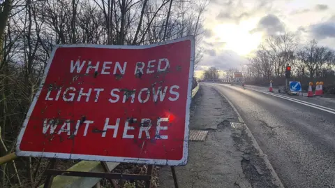 Stuart Woodward/BBC A red sign at the entrance to the bridge, telling motorists to wait at the red light. The bridge has one of its lanes covered with cones and a traffic light.