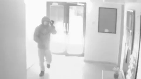 A grab from CCTV footage showing a man running away from the central part of the gym, where its glass doors are showing a fire blazing behind them. The arsonist pictured is wearing a coat, dark shoes and light trousers.