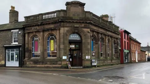 Vikki Irwin/BBC This is a former bank building which has balustrades along the top and large windows which are arched. The building is on a street corner and has a large black door. Above the door is a sign which says "Bank". 