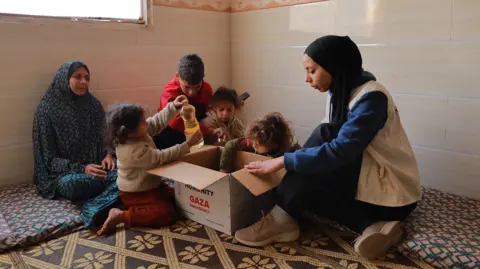Action For Humanity/Fadi Badwan Hanya sits on the floor, opening a cardboard box filled with aid supplies. Four children and another woman look on, two of the children retrieving items from the box.