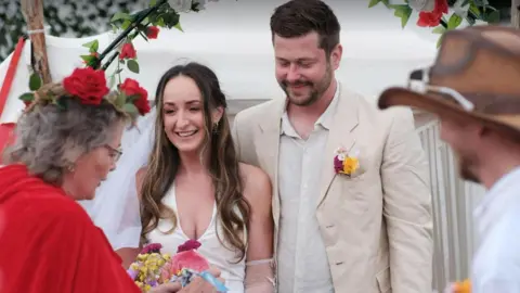 Charlie Lowe and Charles Spires from Harrogate were married in a spiritual hand-tying ceremony in Glastonbury's Healing Field. The wedding will be formally legalised in another ceremony later this year. The celebrant Glenda, left, is wearing a red cloak with red roses in her hair. The bride is wearing a white dress and the groom is in a linen shirt and jacket 