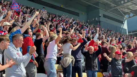 A crowd of happy football fans embrace and cheer, in a football stadium.