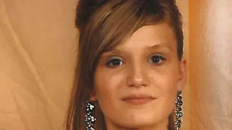 Melissa Mathieson smiles at the camera, she is wearing large shiny blue earrings and makeup, her hair has also been styled and it appears she is going to a formal event.