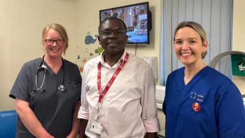 Dr Sian Jenkins (left) with two colleagues at the hospital in Carmarthen. She has grey scrubs on and a stethoscope around her neck. She smiles and has blonde hair and glasses. There is a man in the middle who has a light pink shirt on and a lanyard. He also wears glasses. The woman on the right has blonde hair, which is tied up. She is smiling and has blue scrubs on.