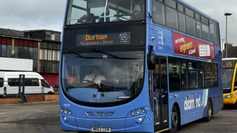 A blue double-decker Go North East bus on the road. The digital sign at the front says it is terminating in Durham.