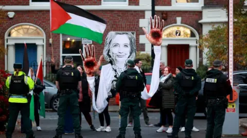 Protestors are pictured standing behind police holding a Palestinian flag and a large black and white cut out of Hilary Clinton with blood on her hands.