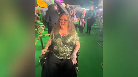 Charlotte Newby Chantelle Nassari sat in a wheelchair. She is wearing a green sparkly top and has blonde curly hair and is wearing glasses. She is at the premiere for the film and there are people standing behind her.