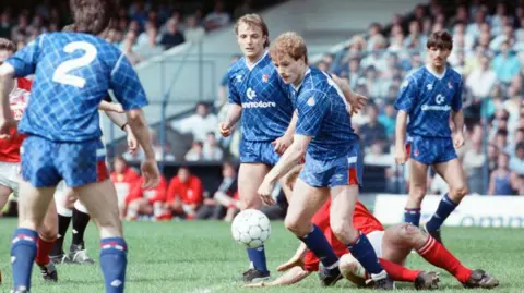 A Chelsea player has control of the ball while a Middlesbrough player attempts to slide tackle him