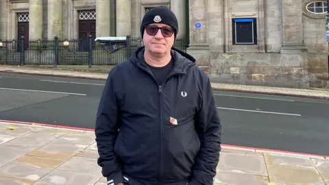 A man wearing a black beanie, sunglasses and a black coat, stands with his hands in his pocket. He is standing on a pavement and behind him across the road is an old, large building.