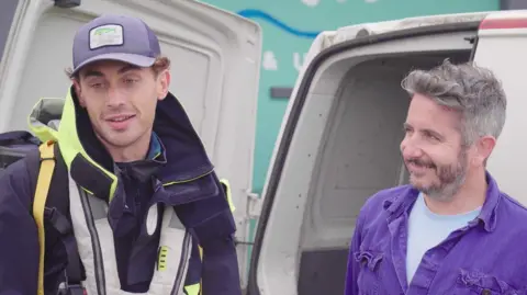 Pain de Minuit - Andoni Martin Marescaux standing next to Steve Oxford who is smiling at Martin. Martin is wearing a dark lifejacket and cap. Steve has a full beard and is wearing a blue shirt.