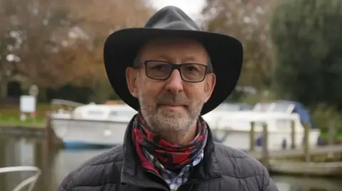 Jamie Niblock/BBC John Packman is standing facing towards the camera. He is wearing a black padded coat, blue and white shirt, red scarf, and a black hat. He has black glasses on and a short grey beard. Behind him is the Broads, with two boats lined up next to each other.